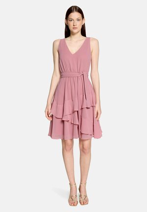 Jeune femme portant une robe rose sans manches à plusieurs couches avec un lien à la taille et des talons dorés à brides, debout devant un fond uni.