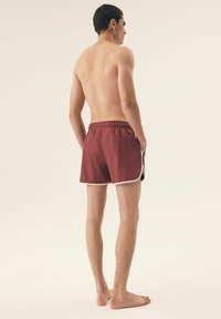 Maroonowe szorty kąpielowe z elastycznym paskiem w talii, białymi akcentami po bokach i luźnym krojem. Widok z tyłu, model jest boso.