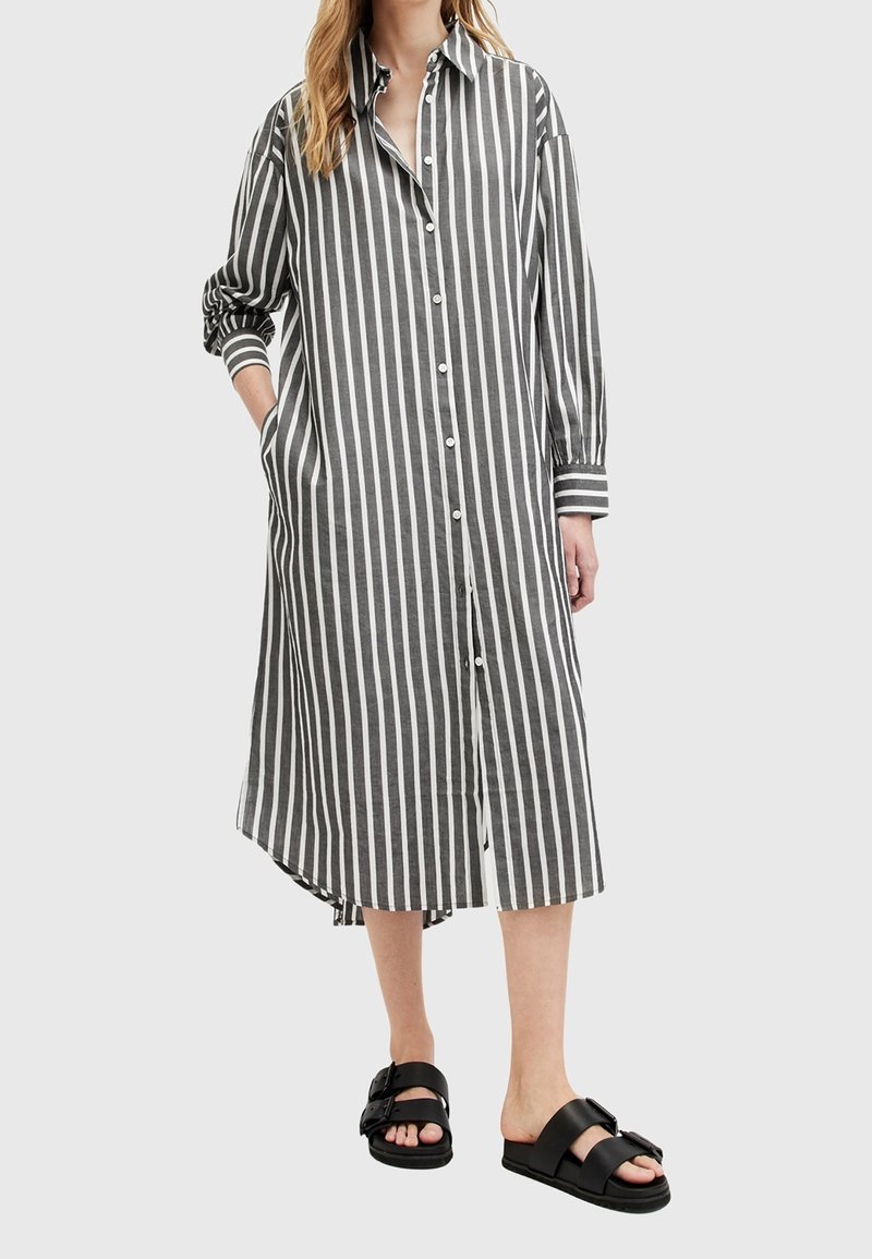 Femme portant une robe longue à boutons à rayures verticales noires et blanches avec des manches longues, et des sandales noires à boucle ouvertes à l'avant.