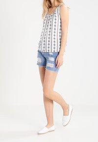White tank top with vertical black geometric pattern, paired with distressed blue denim shorts. White slip-on shoes with woven detail.