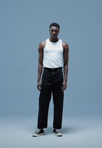 Vit linne, svarta vida byxor och svarta sneakers med hög topp, mot en enfärgad blå bakgrund. Mjukt tyg, avslappnad passform.