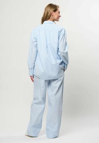 Femme de dos portant une chemise ample bleu clair à fines rayures et un pantalon large assorti sur un fond uni.