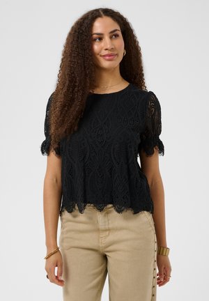 Kvinde med krøllet hår iført en sort blonde kortærmet top og beige bukser, smilende og ser lidt til siden.