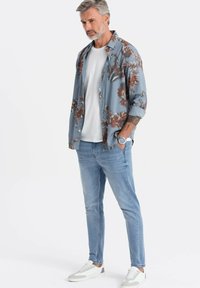 Ljusblå blommig skjorta med knappar över en vit t-shirt, kombinerad med ljusblå jeans och vita sneakers med grå detaljer.