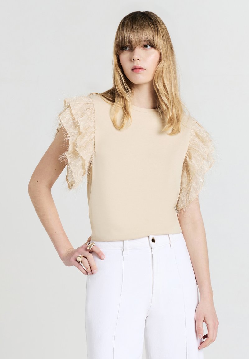 Haut beige avec des manches en tulle texturé et ondulé. Le tissu est lisse, associé à un pantalon blanc ajusté et des bagues aux doigts du modèle.