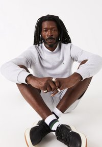 Homme avec des dreadlocks assis en tailleur portant un sweat-shirt gris clair, un short assorti, des baskets noires et des chaussettes blanches à rayures vertes et bleues.