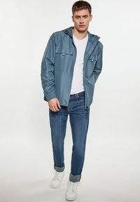 Svetlo modrá nepremokavá bunda s kapucňou, so zipsom a dvoma hrudnými vreckami, skombinovaná s tmavomodrými džínsami a bielymi teniskami.