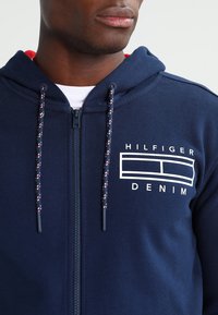 Felpa con cappuccio blu navy con zip, tessuto strutturato, interno rosso e coulisse con motivo. Presenta un logo bianco sul petto.