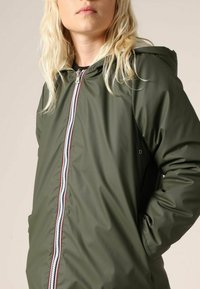 Veste imperméable vert olive avec capuche. Dispose d'une fermeture éclair à l'avant avec un bord contrasté rouge et blanc. Texture lisse et coupe décontractée.