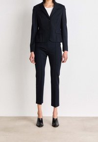Costume sur mesure bleu marine avec un blazer ajusté et un pantalon court. Le blazer est doté d'un bouton unique et de poches avant, accompagné de mules noires à talons.