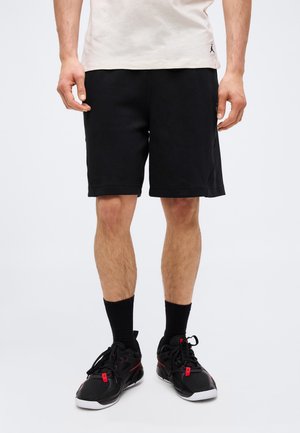 Muškarac u crnim sportskim šortsevima, crnim potkoljenicama, crnim i crvenim tenisicama i bež majici stoji nasuprot jednobojnoj pozadini.