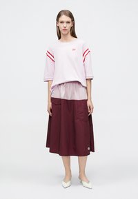 Φαρδιά ροζ φωτεινή μπλούζα με κόκκινες ρίγες και μονόγραμμα; φορεμένη πάνω από μια μεσοφόρια μαρόνη φούστα με υφή και τσέπες. Λευκά παπούτσια.