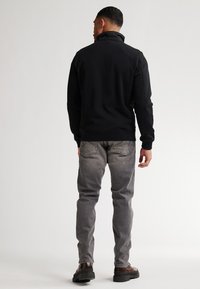Chaqueta de forro polar negra con cuello alto, combinada con jeans grises. Las características incluyen una textura suave, un diseño ajustado y zapatos marrones resistentes.