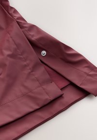 Detail vínovočervenej nepremokavej bundy s hladkou textúrou, strieborným patentným zapínaním a starostlivo prešitými okrajmi, zobrazujúci vnútornú podšívku.