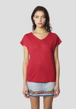 Rdeča kratka majica z V-izrezom in lahko teksturo, z nežno vezenim logotipom. Nošena z denim krilom, okrašena z rožnim vezenjem.