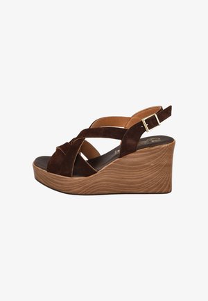 Sandalia cuña de ante marrón con tiras cruzadas y una hebilla dorada sobre un tacón plataforma con textura de madera sobre fondo blanco.