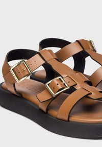 Sandalias de cuero marrón que presentan dos correas anchas, hebillas cuadradas doradas y una suela negra gruesa para mayor comodidad y estabilidad.