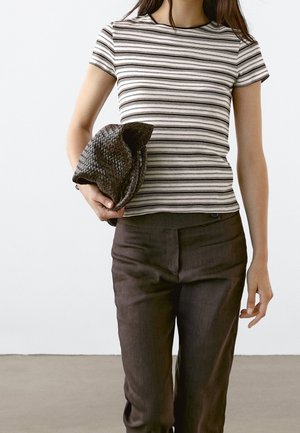 Striped short-sleeve top in neutral tones paired with dark brown tailored pants. Model holds a woven black hat in her left hand.