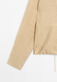 Beige long-sleeve shirt with a visible left sleeve, side seam pocket, and drawstring hem on a white background.