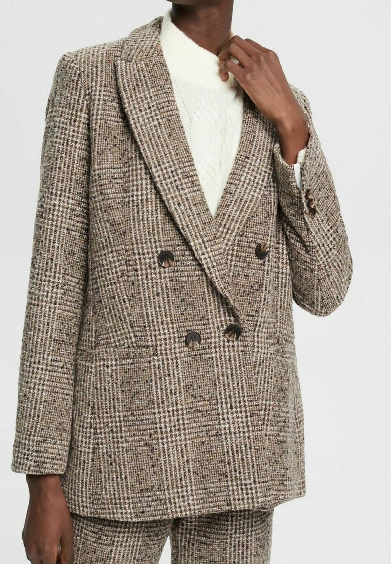 Personne portant un blazer croisé à carreaux marron et beige et un pantalon assorti avec un pull blanc texturé en dessous.