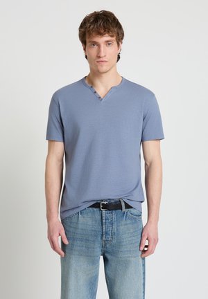 Giovane uomo in piedi che indossa una maglietta henley a maniche corte di colore azzurro chiaro infilata in jeans blu slavati con una cintura nera, espressione neutra.