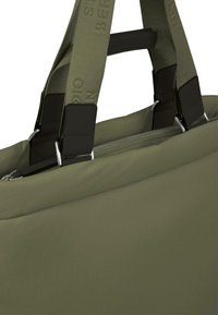 Olivengrønt taske med robust stof, lynlåslukning og forstærkede sorte håndtag. Har brede stropper med prægede brandingdetaljer.