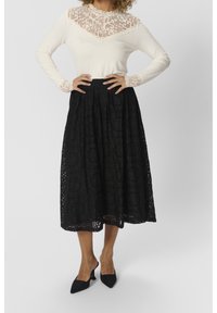 Crème top met lange mouwen en kantdetails, gecombineerd met een zwarte kanten midi-rok met bloemmotieven. Zwarte puntige hakken maken de look af.