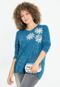 Maglione teal con motivo di fiocchi di neve argentati e dettagli luccicanti. Dotato di maniche lunghe e scollatura rotonda. Abbinato a una borsa argentata.