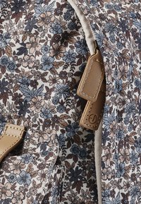 Blommönstrad tyg i dämpade toner av blått, beige och brunt. Har en krämfärgad kant och beigea läderdetaljer med stickade detaljer.