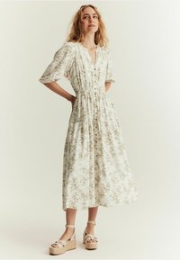 Vitt blommigt midiklänning med korta puffärmar, V-ringning, knappdetaljer och elastisk midja. Matchas med beige kilklackar.