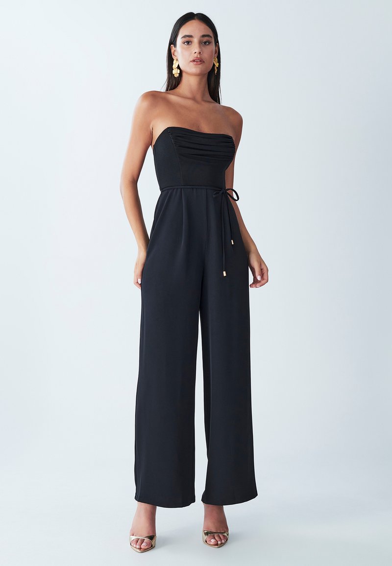 Zwarte strapless jumpsuit met een getailleerd lijfje, textuurstof, wijd uitlopende broekspijpen en een trekkoord in de taille. Gouden accenten op de schoenen.