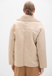 Giacca in finta suede beige con colletto in morbido shearling, forma oversize e dettagli di cuciture visibili, mostrata da dietro.