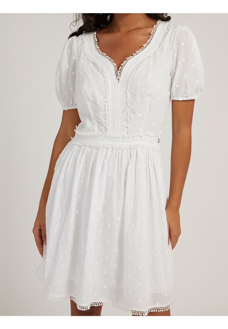 Femme portant une robe blanche mi-longue aux manches bouffantes, à pois texturés, ornée de dentelle et taille froncée.