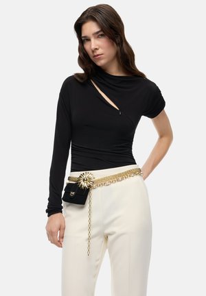 Mujer con top negro asimétrico de manga larga, pantalones blancos y cinturón de cadena dorada con detalle de flor y pequeño bolso negro.