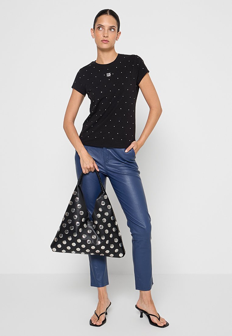 Femme portant un t-shirt noir à clous, un pantalon en cuir bleu, des sandales à talons noires, tenant un grand sac à main noir à pois blancs, se tenant devant un fond blanc.