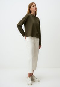 Maglietta verde oliva a maniche lunghe, vestibilità ampia, tessuto testurizzato, abbinata a pantaloni bianchi a gamba dritta e sneakers bianche su uno sfondo neutro.