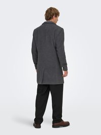 Manteau gris en laine avec une coupe ajustée, revers crantés, fente unique au dos, accompagné d'un pantalon noir et de chaussures marron.