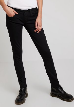 Osoba stojící s jednou rukou v kapse, oblečená v černých úzkých džínách a černých šněrovacích botách, na jednobarevném pozadí.