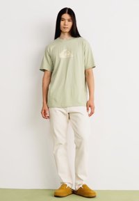 Light green cotton t-shirt with a small white logo, paired with white straight-leg pants and brown shoes. Simple, casual design.