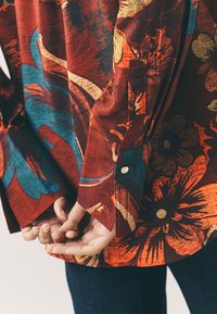 Red patterned shirt with blue and yellow floral designs, featuring long sleeves and button cuffs. Fabric has a textured appearance.