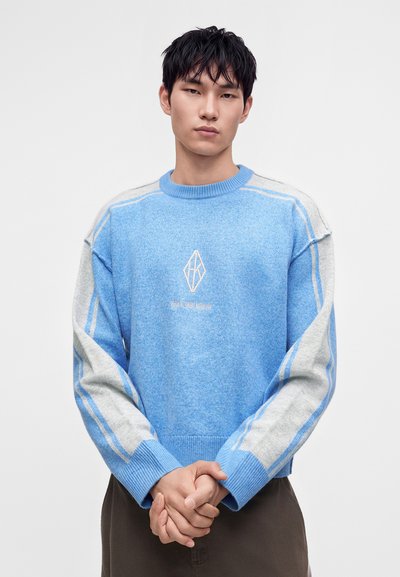 Jeune homme aux cheveux courts noirs portant un pull bleu et gris à manches longues avec un logo géométrique, debout les mains jointes devant lui.