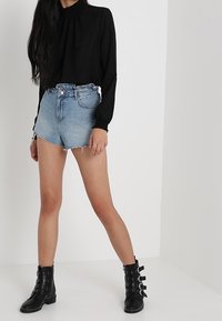 Blouse noire à manches longues avec un col smocké, associée à un short en denim déchiré et des bottines noires avec boucles et accents cloutés.