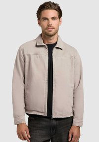 Homme aux cheveux bruns courts portant une veste zippée beige clair par-dessus un t-shirt noir, debout devant un fond gris uni.