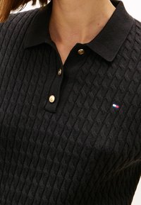 Gros plan d'un polo en maille noire avec un motif torsadé, des boutons dorés et un petit logo rectangulaire rouge, blanc et bleu sur la poitrine.