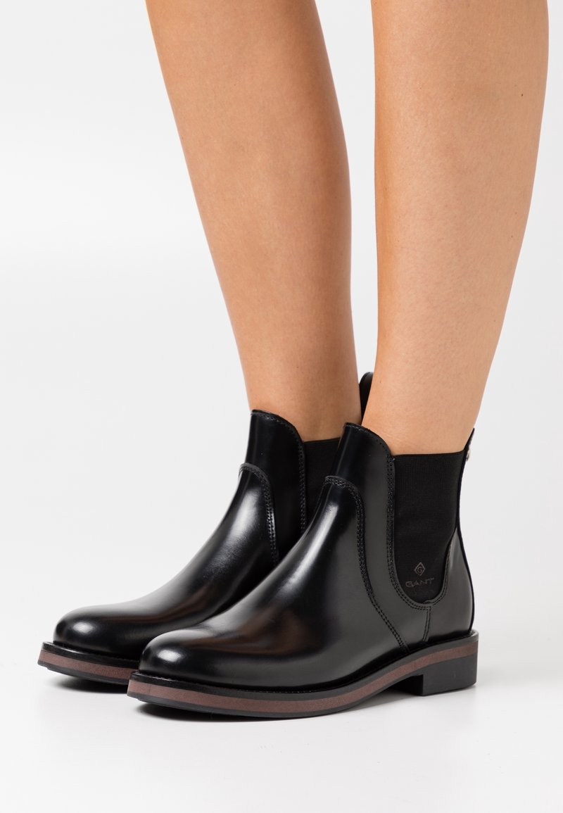 Bottes chevilles en cuir noir avec panneaux élastiques sur les côtés et talons bas, portées sur des jambes nues, photographiées sur un fond blanc.