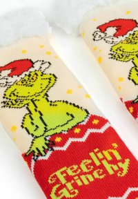Calzini rossi e crema con una grafica del Grinch verde che indossa un cappello di Babbo Natale. Il testo recita "Me ne sento un po' Grinch" con accenti gialli e una morbida cima bianca.