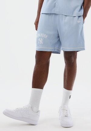 Man draagt lichtblauwe New York Yankees-short, witte sportsokken en witte Nike-sneakers, staand tegen een effen witte achtergrond.