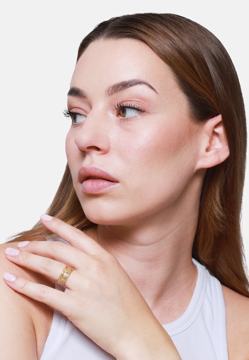 Goldring an einer Hand mit langen Fingern, glatter Haut und zart rosa Nägeln. Der Hintergrund ist schlicht weiß, was den Ring betont.