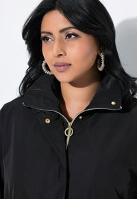 Svart zip-up-kappa med hög krage, dekorerad med guldfärgad hårdvara och en metallisk dragkedja, visad på en person som bär hoops örhängen.