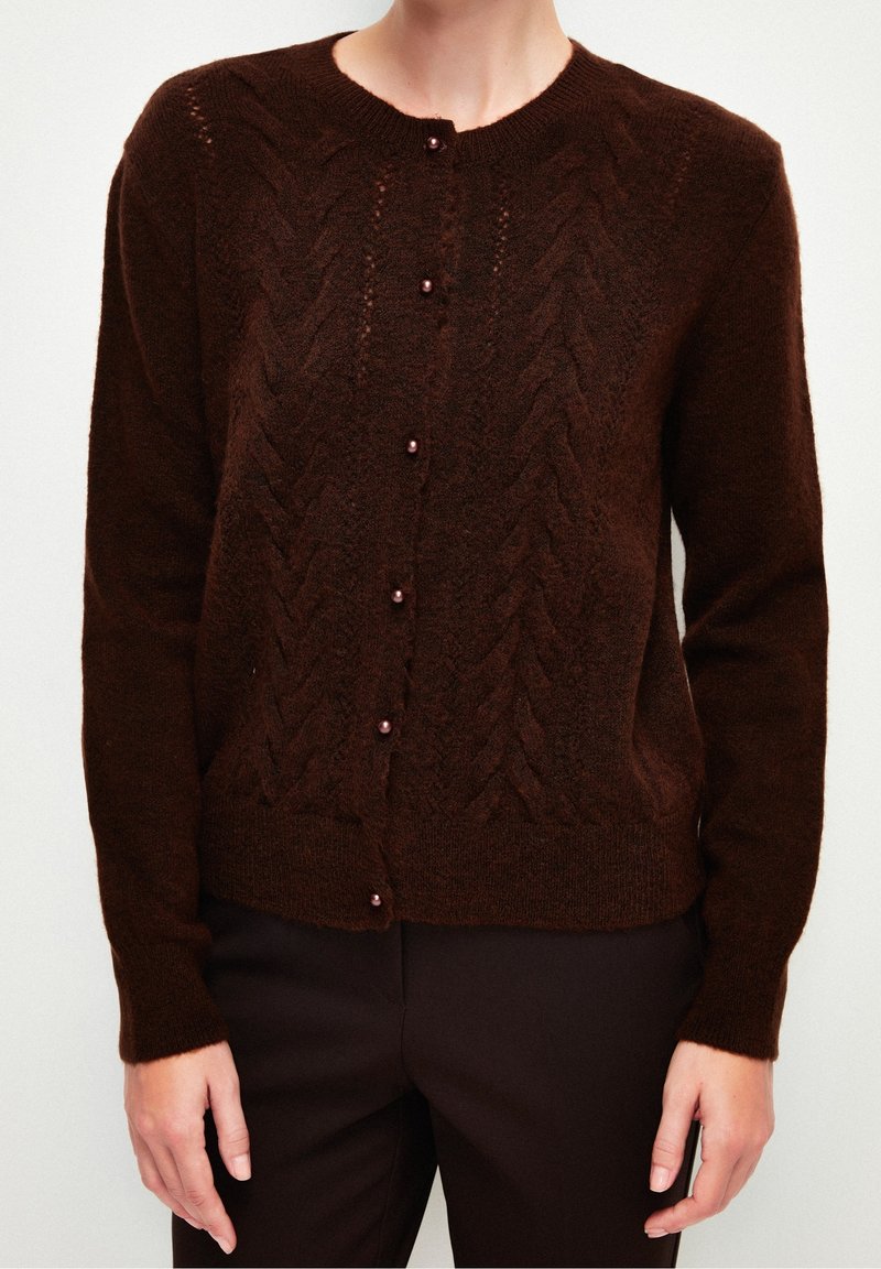 Cardigan en tricot marron avec un motif en chevrons texturé, fermeture à boutons avec des boutons ronds et brillants, et des poignets et un ourlet côtelés.